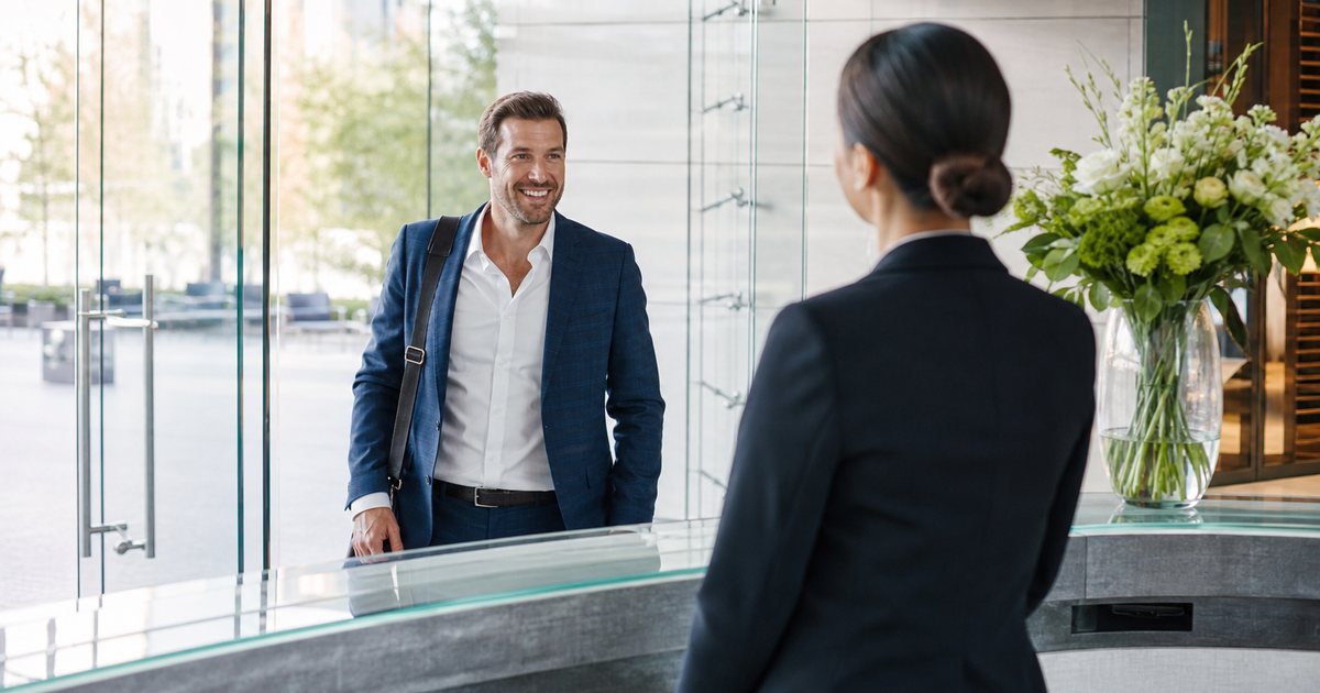 A well-dressed business guest arriving at a bright modern office lobby, a receptionist standing to greet them, glass doors and morning light, fresh flowers on the desk
